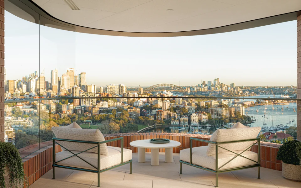 A luxury balcony with two modern armchairs overlooking a panoramic view of the Sydney Harbour Bridge, Opera House, and city skyline at sunset.