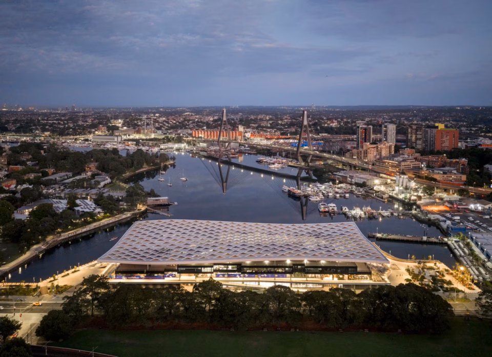 Aerial twilight view of the new Sydney Fish Market featuring a distinctive white patterned roof, situated on Blackwattle Bay with the illuminated Anzac Bridge in the background.