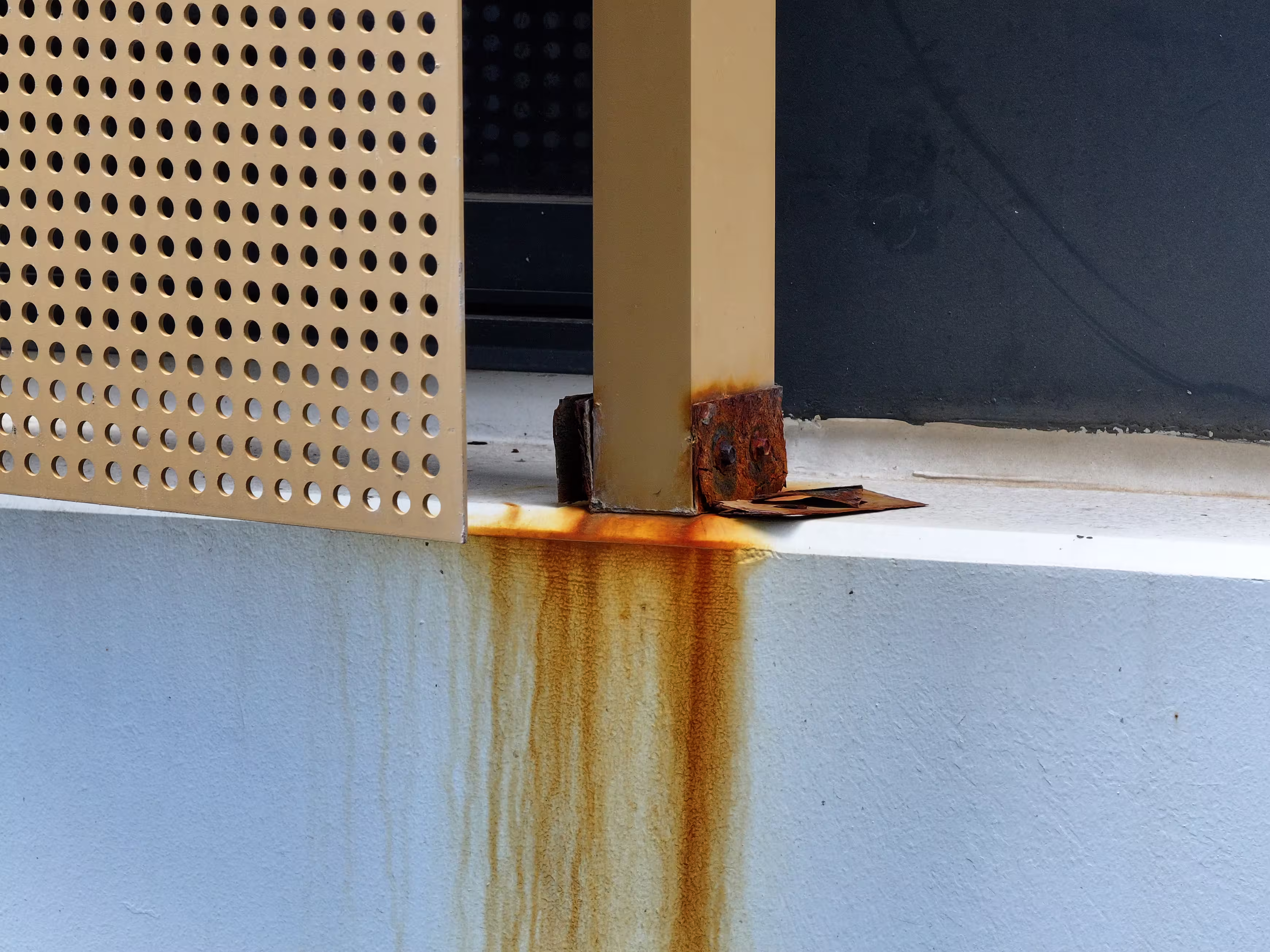 A close-up inspection photograph showing heavy orange rust corrosion and staining on a metal support beam and perforated panel attached to a white concrete ledge.