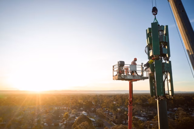 Workers in a high-reach platform use a crane to position a large green cellular antenna array onto a telecommunications tower at sunset in Greater Sydney.