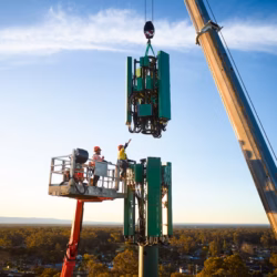 Workers in a high-reach platform use a crane to install a large green cellular antenna array onto a telecommunications tower over Greater Sydney.