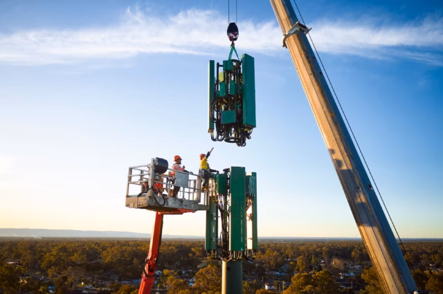 Workers in a high-reach platform use a crane to install a large green cellular antenna array onto a telecommunications tower over Greater Sydney.