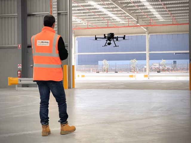 A technician in an orange SkyMonkey safety vest monitors a large industrial drone hovering at a warehouse entrance.