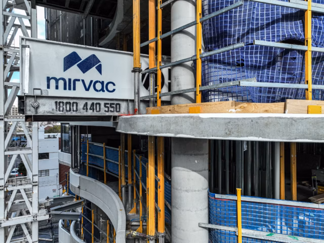 A close-up view of a Mirvac construction site featuring a branded structural beam, concrete pillars, and blue safety netting on a multi-story building.