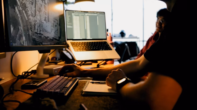 A dark, moody shot of a production workstation where an editor uses a mechanical keyboard and dual monitors to review aerial maps and project data